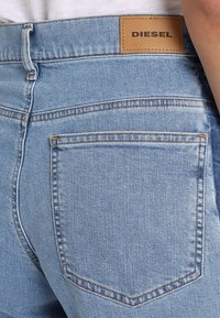 Světle modré džíny z denimu s hnědou koženou nášivkou s označením "DIESEL" na zadní straně. Standardní design kapes s viditelným šitím.