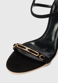 Black suede heeled sandal featuring a gold hardware accent with a crystal detail, showcasing a sleek design and open-toe style.