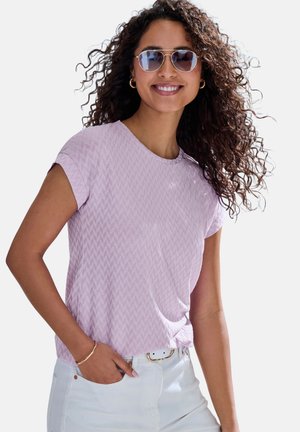 Hellviolettes T-Shirt mit kurzen Ärmeln und einem strukturierten Chevron-Muster, rundem Halsausschnitt, getragen mit weißen Hosen und mit einer Sonnenbrille als Accessoire.