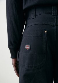 Pantalon en denim noir avec une taille ajustée, présentant un patch logo Harley-Davidson bien visible et des accents en métal doré sur les poches.