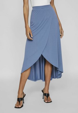 Femme portant une jupe portefeuille bleue asymétrique avec un ourlet haut-bas et des sandales noires à talons ouverts.