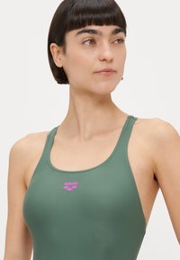 Bañador verde hecho de material suave, con diseño de espalda cruzada y un pequeño logo rosa en el pecho.