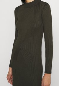 Mörk olivgrön ribbad stickad klänning med långa ärmar och hög krage, bärs av en person som är sedd från axlarna till mitten av låret.