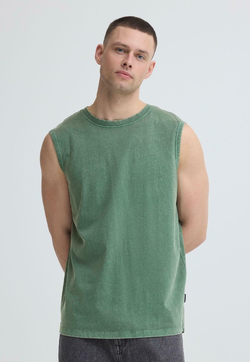 Νέος άνδρας με κοντά μαλλιά, φορώντας ένα μουντό πράσινο αμάνικο πουκάμισο, στέκεται μπροστά σε έναν απλό ανοιχτό γκρι φόντο με τα χέρια πίσω από την πλάτη.