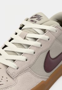 Gros plan d'une basket beige Nike SB Force 58 avec des lacets blancs, un swoosh Nike bordeaux et une semelle couleur gomme.