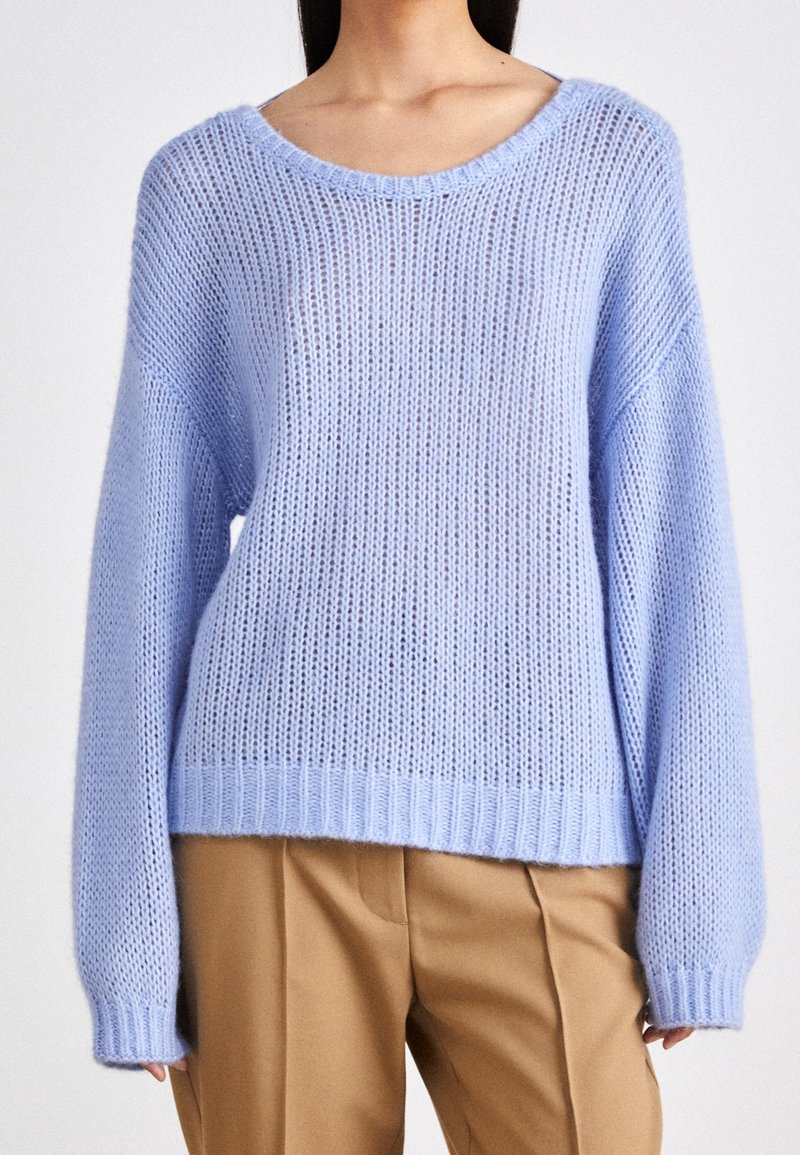Ljusblå stickad tröja med lös passform, bred halsringning och långa ärmar. Materialet har en strukturerad, öppen vävdesign. Kombinerad med beiga byxor.