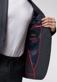 Grau kariertes Sakko mit blauer Innenauskleidung und roten Akzenten. Das Innere verfügt über Etiketten mit der Aufschrift "SLIM FIT" und "Stretch". Getragen über einem weißen T-Shirt.