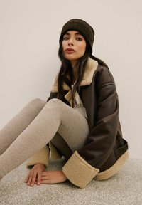 Brązowa skórzana kurtka z beżowym kołnierzem i mankietami z owczej wełny, w zestawie z jasnobrązowymi legginsami o teksturowanej powierzchni oraz ciemnym dzianinowym czapek.