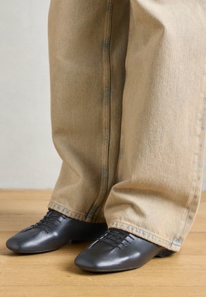 Črne usnjene čevlje z gladko teksturo, nizkim profilom in vezalkami, v kombinaciji z ohlapnimi, svetlo bež traper hlačami.