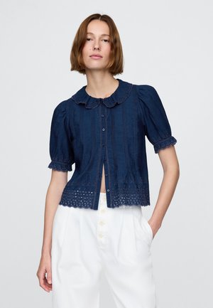 Meresinine pluus lühikeste pufvarrukatega, lainelise krae ja silmaaugulise pitsdetailiga ääres. Esiküljel on nööbitav ja kangas tekstuuriga.