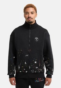 Black zip-up sweatshirt with a high collar; features colorful splatter patterns at the bottom and a logo on the chest. Soft fabric, relaxed fit.