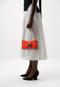 Sac à main en cuir rouge avec une forme structurée et du matériel argenté, porté à la main. La tenue comprend une jupe blanche et des escarpins noirs.