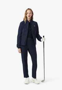 Granatowa pikowana kurtka z wysokim kołnierzem, połączona z dopasowanym polo i eleganckimi spodniami. W komplecie kij golfowy i białe buty.