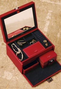Caja de joyería de cuero rojo con tapa espejo, interior de terciopelo azul, compartimentos para accesorios y dos cajones anidados. Acentos dorados visibles.