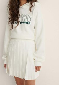 Valkoinen huppari, jossa on vihreällä teksti "CLUB OFFICE", yhdistettynä pleatattuun valkoiseen mini-hameeseen. Kangas näyttää pehmeältä ja sileältä.