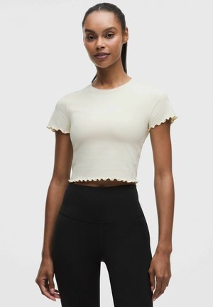 Kvinde iført en tætsiddende cropped cremefarvet t-shirt med flæsekant og højtaljede sorte leggings, stående mod en ensfarvet baggrund.