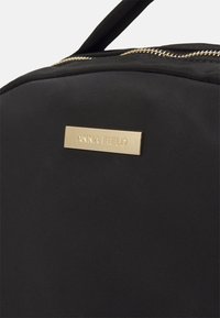 Bolso de nylon negro con una placa de metal en tono dorado con la inscripción "ANNA FIELD" grabada. Tiene una textura suave y un cierre con cremallera.