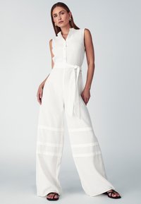 Mono de lino blanco con diseño sin mangas, cierre de botones, silueta de pierna ancha, cinta en la cintura y detalles de rayas horizontales.