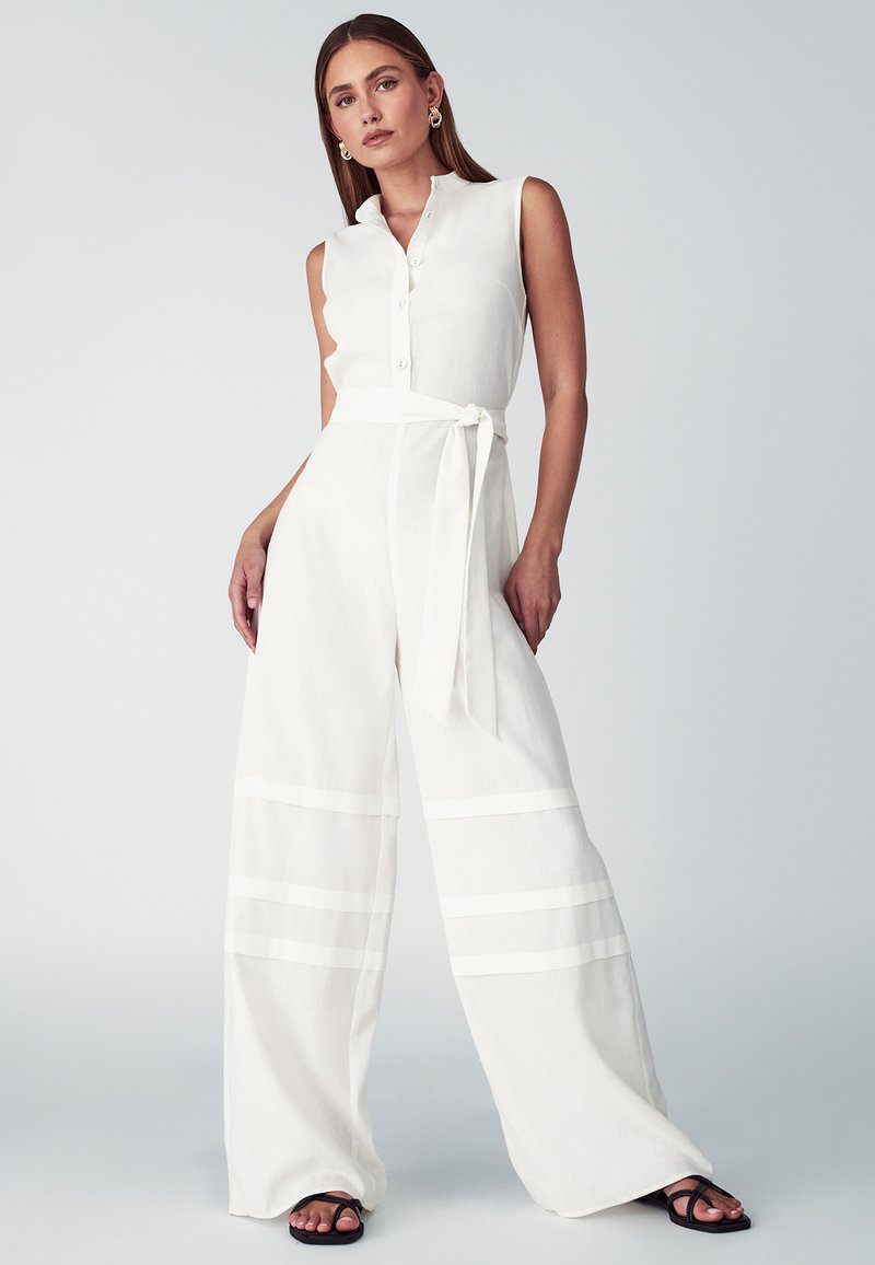 Mono de lino blanco con diseño sin mangas, cierre de botones, silueta de pierna ancha, cinta en la cintura y detalles de rayas horizontales.