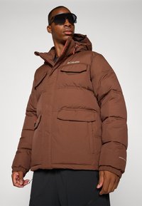 Braune Pufferjacke mit Kapuze, ausgestattet mit einem Reißverschluss vorne, vier externen Taschen und elastischen Bündchen. Logo auf der Brust und Kordelzüge an der Kapuze.