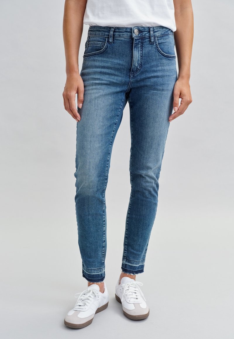 Jean bleu slim avec ourlets effilochés, porté avec des baskets blanches et un haut blanc, montré de la taille aux pieds sur un fond neutre.