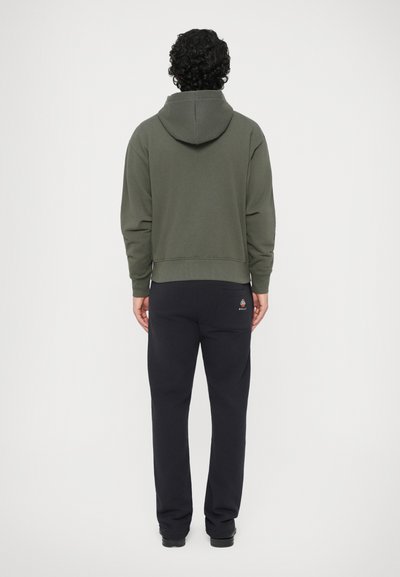 Persona con cabello rizado de pie de espaldas, vistiendo una sudadera verde olivo y pantalones negros con un pequeño logo colorido en el bolsillo trasero.
