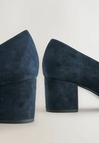 Zapatos de tacón alto de ante azul marino con un tacón grueso, con punta afilada y textura suave, que capturan un diseño minimalista.