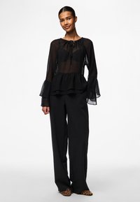Black sheer blouse with ruffled peplum, long puffed sleeves, and a tie neckline. Paired with black wide-legged trousers and patterned shoes.