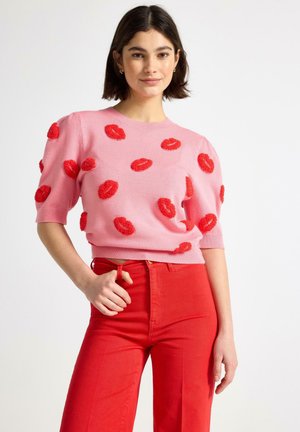 Nainen pukeutuneena vaaleanpunaiseen lyhythihaiseen neuleeseen, jossa on punahuulinen kuosi, ja korkeavyötäröisiin punaisiin housuihin, seisomassa yhdellä kädellä taskussa.