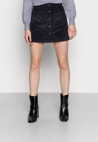 Námořnicky modrá manšestrová minisukně se zapínáním na knoflíky vpředu, dvěma předními kapsami a přiléhavým střihem. Kombinováno s černými kotníkovými botami.