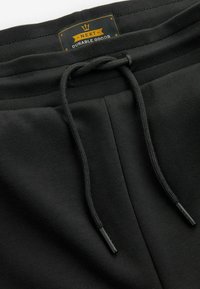Černé jogger kalhoty s elastickým pasem na stažení, které mají texturovaný elastický pás a štítek s nápisem "NEXT DURABLE GOODS." Měkká tkanina.