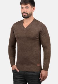 Brun v-hals sweater lavet af blødt strikket stof, med ribkant i talje og ærmer, samt et lille lædermærke i bunden.