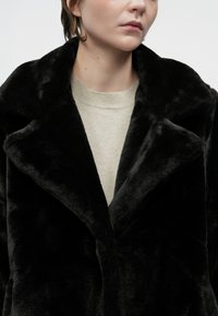 Pessoa vestindo um casaco de pelo sintético preto sobre um suéter creme claro de gola redonda, mostrada do queixo até a metade do torso contra um fundo simples.