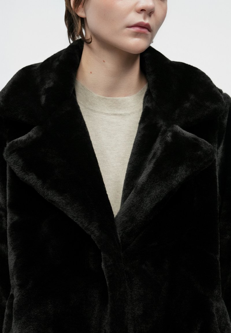 Person trägt einen schwarzen Kunstpelzmantel über einem hellbeigen Rundhalsausschnitt-Pullover, gezeigt von Kinn bis zur Mitte des Oberkörpers vor einem schlichten Hintergrund.