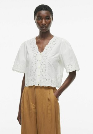 Weiße Baumwollbluse mit kurzen Puffärmeln, V-Ausschnitt und Lochstickerei entlang der Vorderseite und dem Saum. Kombiniert mit beigen weiten Hosen.