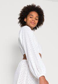 Top crop de algodão branco com mangas balão, apresentando detalhes de ilhós bordados e uma cintura justa. Textura suave e design arejado.