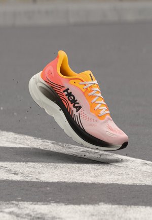 Chaussure de course Hoka orange vif et rose en l'air au-dessus de l'asphalte avec des marquages routiers blancs, montrant une semelle noire et un logo sur le côté.
