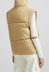 Beige vadderad väst med hög krage och ärmlös design, med en mjuk textur. Matchas med en långärmad vit topp med utskärningsdetalj.