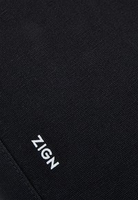 Czarna teksturowana tkanina z białą nazwą marki "ZIGN" wydrukowaną blisko obszytego brzegu.