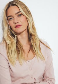 Gaiši rozā džemperis, kas izgatavots no mīksta dzijas, ar apaļu kakla izgriezumu un mazām dekoratīvām pogām. Garie, viļņainie mati ietver seju.