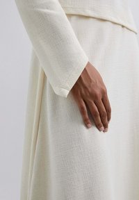 Indumento bianco con texture, caratterizzato da maniche lunghe e un discreto motivo a intreccio, con una mano appoggiata sul tessuto. Forma morbida e drappeggiata.