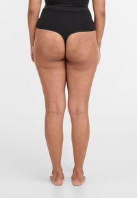 Vrouw die blote voeten heeft, gekleed in zwarte hoog-taille tangashorts, gezien vanuit de achterkant tegen een effen witte achtergrond.