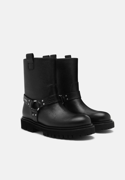 Stivaletti neri in pelle con punta tonda, suola spessa e una fascia con accessori metallici. Finitura testurizzata e linguette di estrazione nella parte superiore.