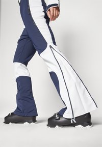 Tumesinised ja valged suusapüksid, mille külgedel on tõmblukud, koos mustade suuskadega, millel on valged aktsendid ja reguleeritav rihm.