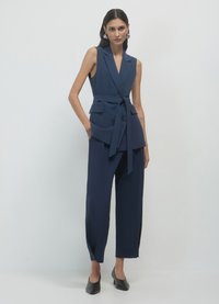 Navyblauer taillierter Weste mit Wickeldesign, gerafftem Taillenzug und zwei Fronttaschen, kombiniert mit locker sitzenden Hosen und schwarzen Absätzen.