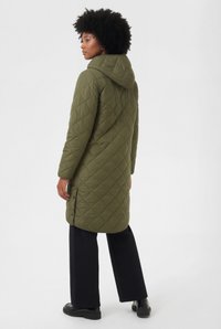 Steppmantel in Olivgrün mit Kapuze, mit Rautenmuster, seitlichen Knöpfen und geradem Schnitt, getragen über schwarzer Hose und Stiefeln.