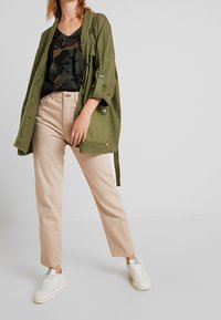 Olivgrön lättviktsjacka över en svart kamouflage t-shirt, tillsammans med beige raka byxor och vita sneakers. Texturerade tygdetaljer synliga.