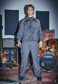 Eine Person mit Tätowierungen, die einen grauen Trainingsanzug trägt, steht in einem Musikstudio, umgeben von Gitarren, Verstärkern und einem Yamaha Schlagzeug.