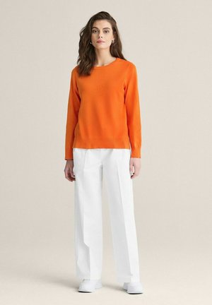 Orange langærmet sweater med rund hals, parret med hvide vide bukser og hvide sko. Simpel, glat tekstur, afslappet pasform.