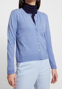 Světlomodrý kardigan s knoflíkovou légou vpředu, žebrovaným lemem a dlouhými rukávy. Nosit přes přiléhavý tmavě modrý rolák, vytvářející vrstvený vzhled.